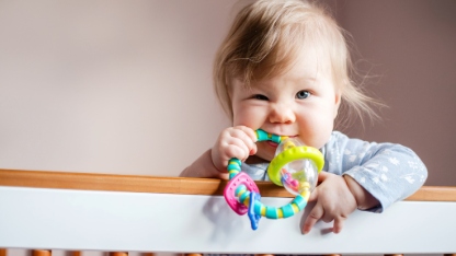 Cute Caucasian little baby girl standing in her crib and chewing on an infant toy, looking at the camera, with copy space. 