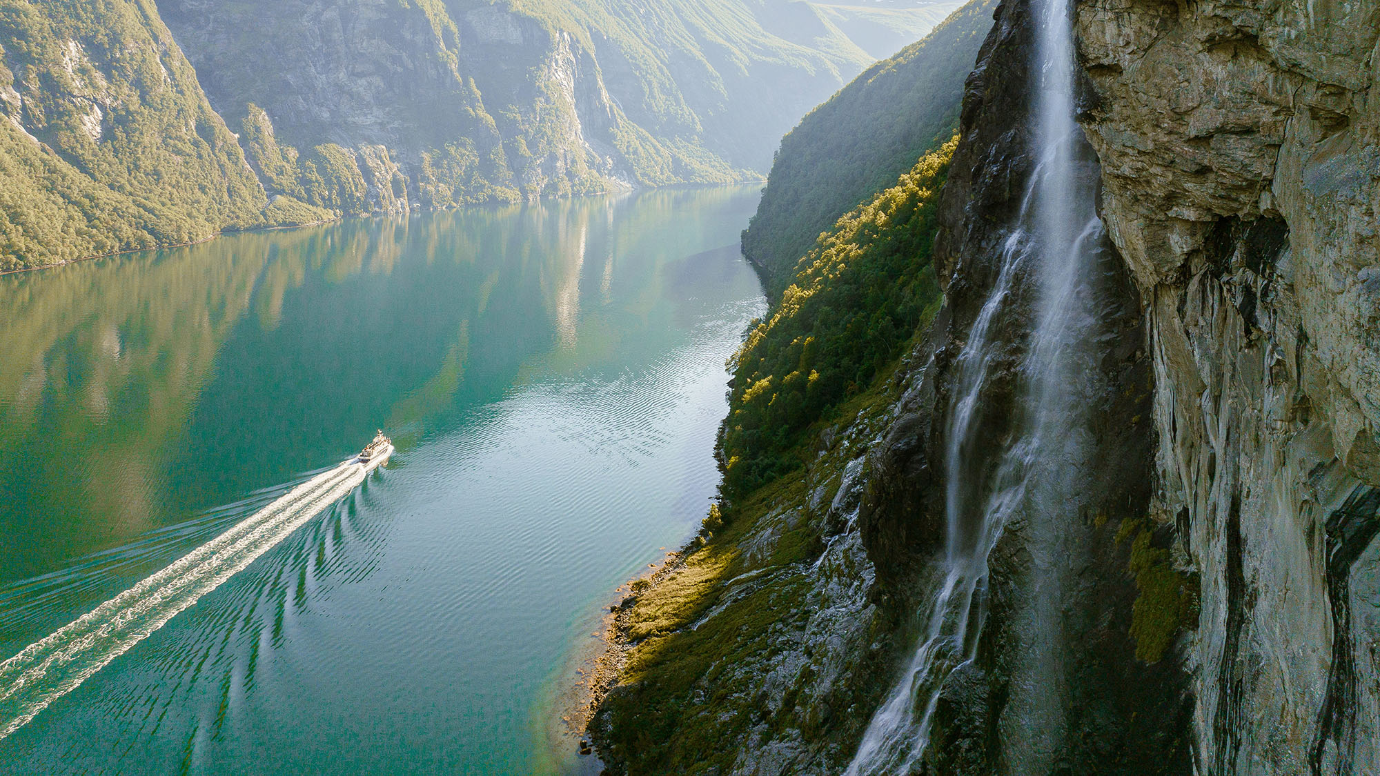 Ship on the water, passing through a canyon with a waterfall and mountains in view 